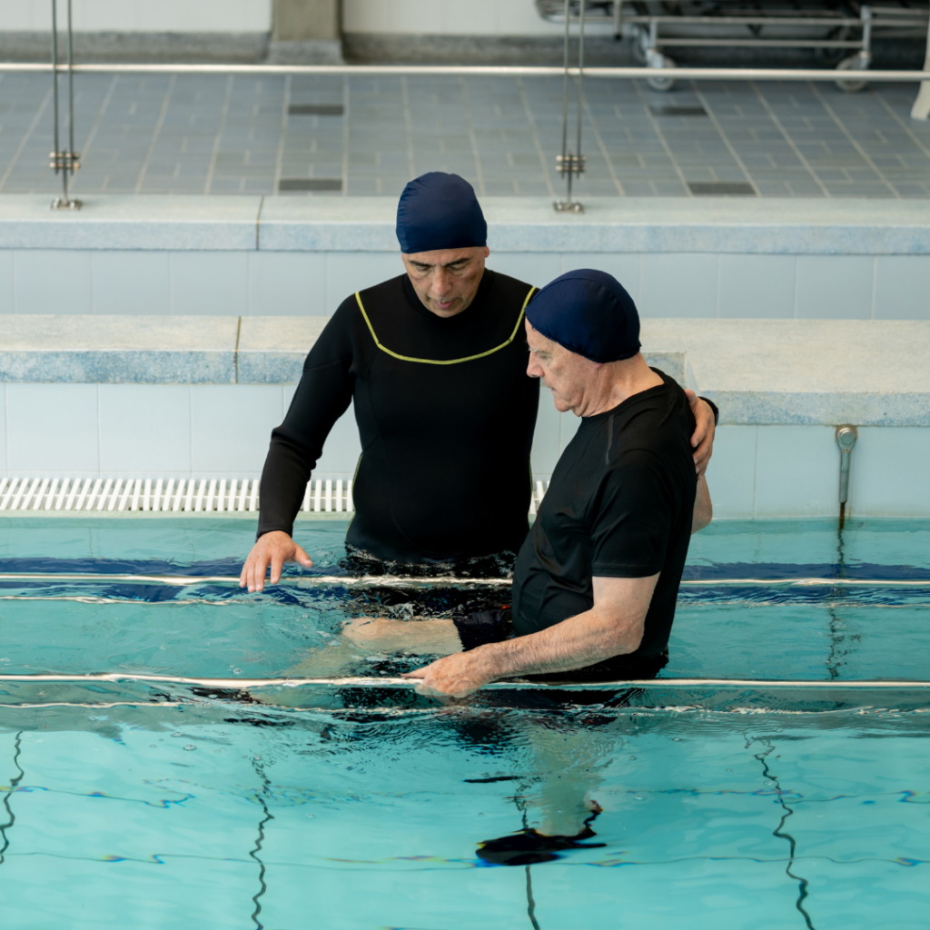 An older man undergoing an NDIS hydrotherapy session with a physiotherapist assisting him in a warm indoor pool, focusing on gentle movement and rehabilitation exercises.