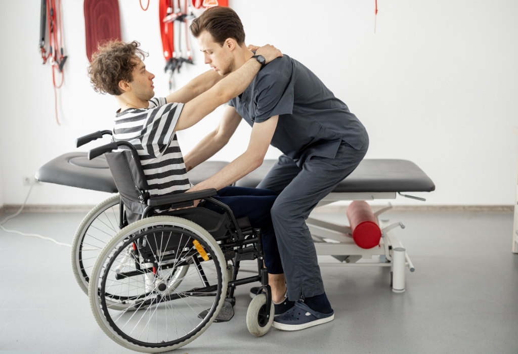 A physiotherapist providing support to help a person in a wheelchair stand up, showcasing personalised care and rehabilitation through mobile physiotherapy services.