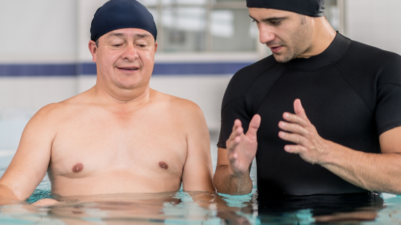 A therapist explains hydrotherapy exercises for fibromyalgia for to a man with a disability in a swimming pool, demonstrating the benefits of hydrotherapy for arthritis and joint mobility.