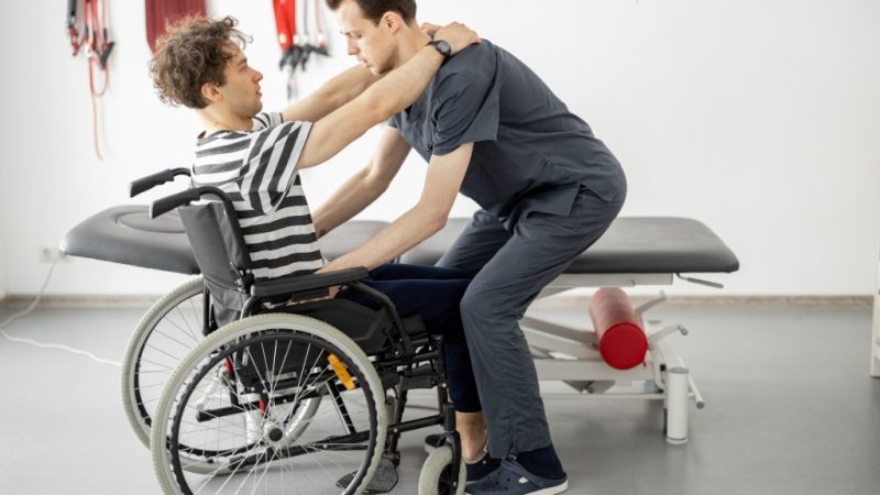 A physiotherapist providing support to help a person in a wheelchair stand up, showcasing personalised care and rehabilitation through mobile physiotherapy services.